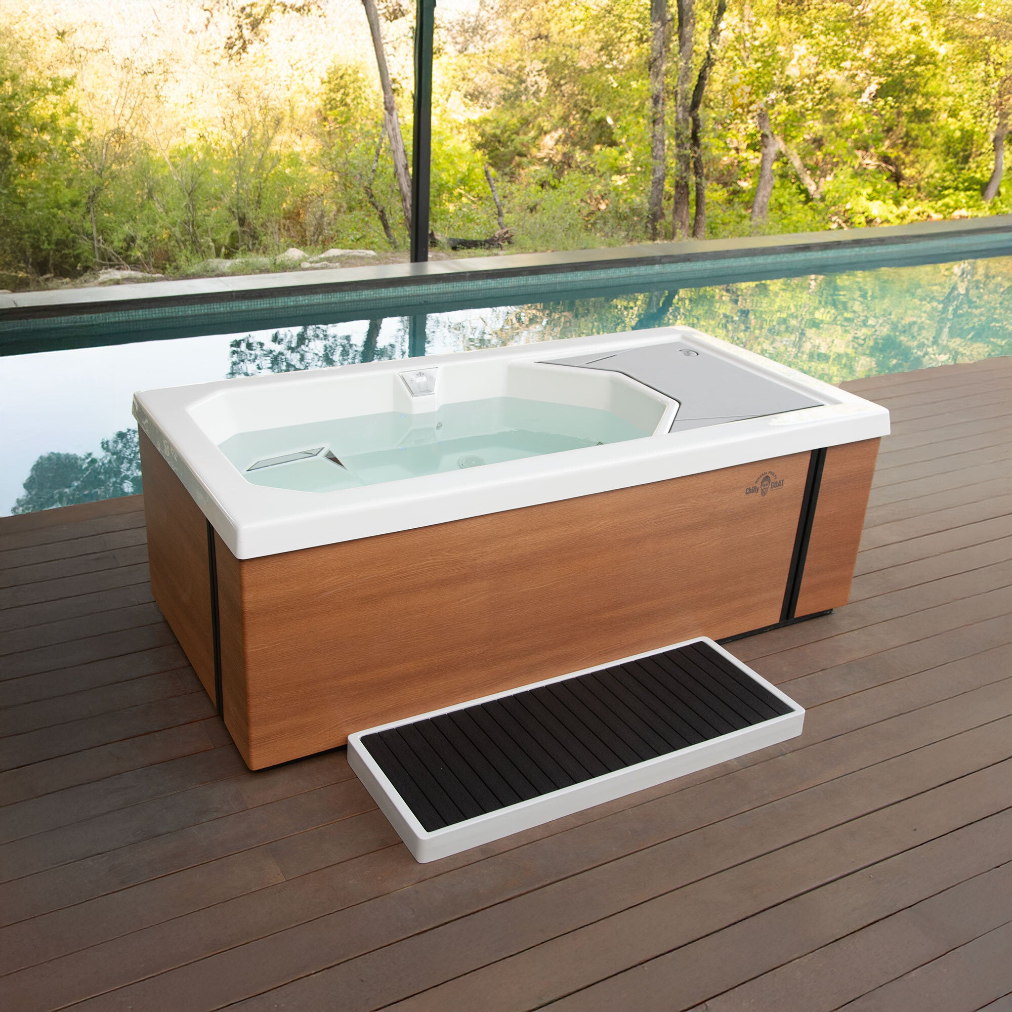 Hot tub with wooden exterior and white interior on a wooden deck with a pool and forest in the background.